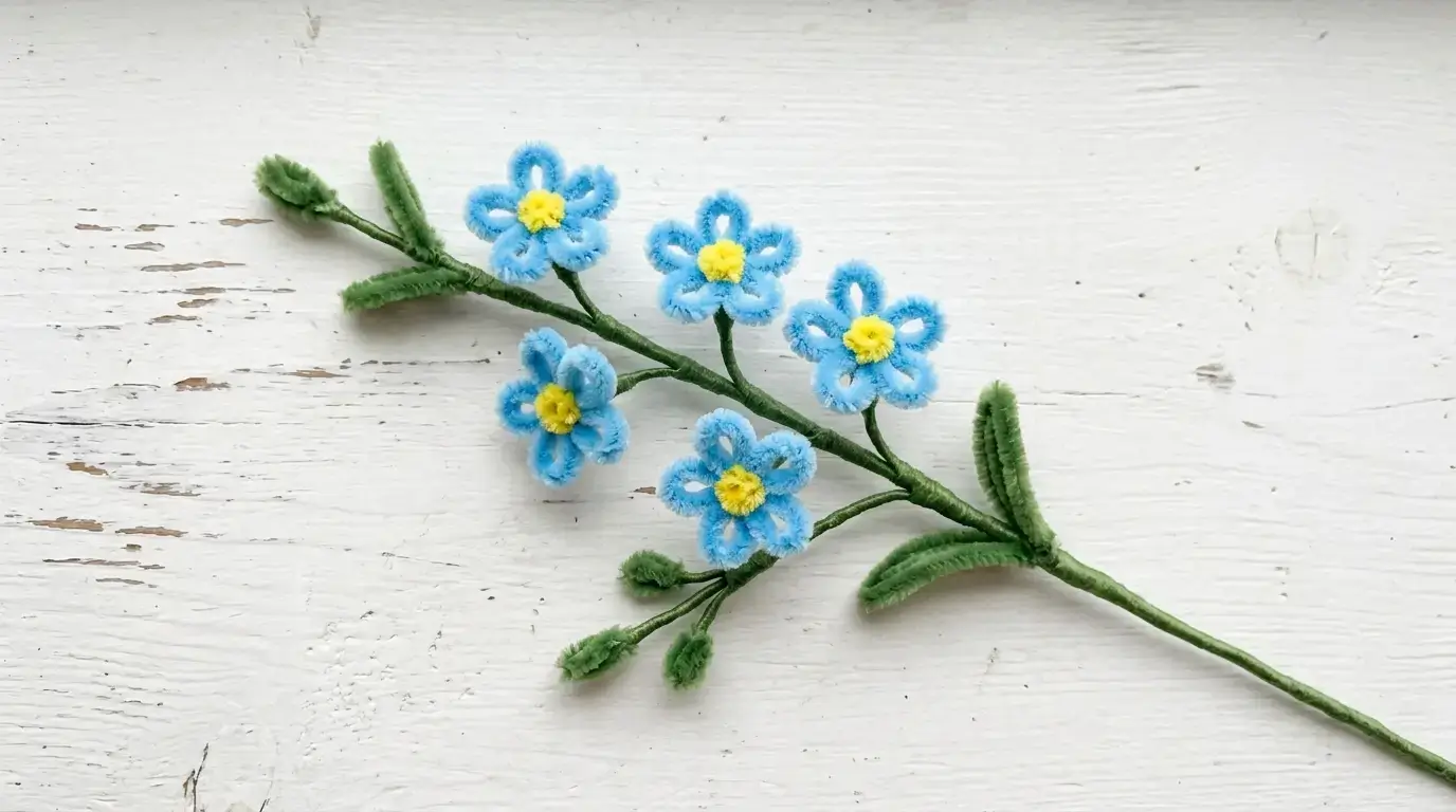 Handmade pipe cleaner forget-me-not spray with tiny blue petals and yellow centers on a green stem displayed on a white surface