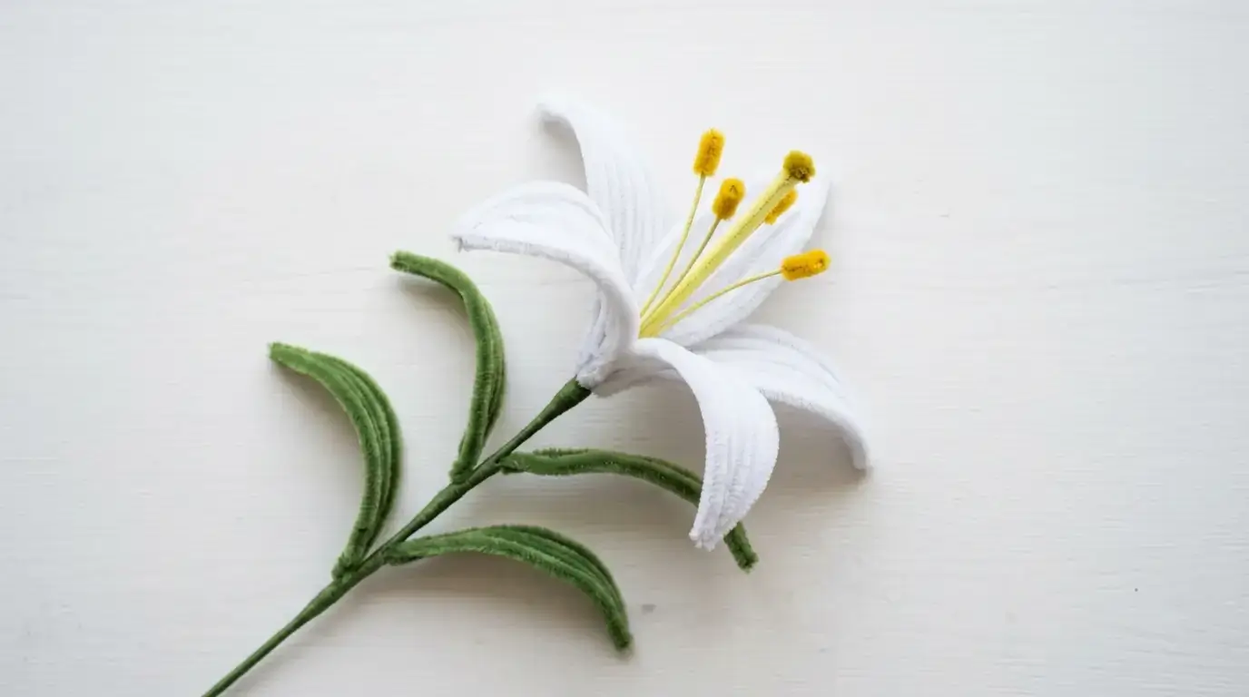 Handmade pipe cleaner lily with white petals and yellow stamens displayed on a white surface next to green leaves